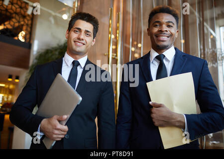 Confiant joyeux partenaires commerciaux multi-ethnique en costumes officiels debout dans un hall moderne et looking at camera, ils holding laptop et documents Banque D'Images