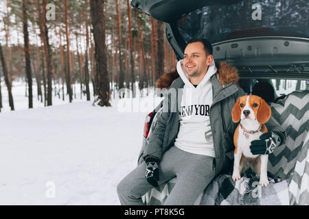 Un jeune beau mec dans une forêt d'hiver neigeux est assis dans le coffre de sa voiture dans une étreinte avec son ami chien beagle. Promenez le chien Banque D'Images