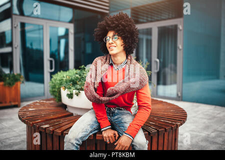 Les jeunes habillés à la mode afro-américain noirs bouclés avec des lunettes homme est assis sur un banc dans la rue près de la vitre des murs du bâtiment. Un étudiant Banque D'Images