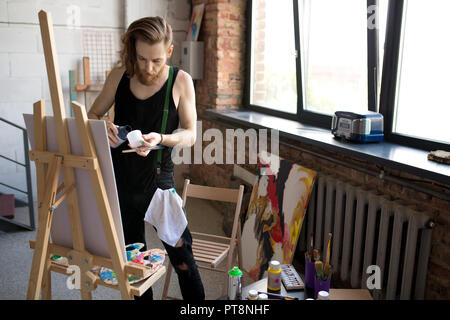 Portrait de l'artiste aux cheveux longs contemporain mélange des peintures tout en travaillant par chevalet dans art studio, copy space Banque D'Images