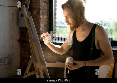 Vue de côté portrait de l'artiste aux cheveux long porté la tenue du pinceau et de la peinture sur chevalet dans art studio éclairée par la lumière du soleil, copy space Banque D'Images