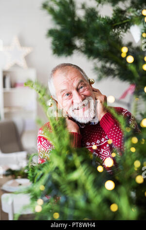 A senior man standing by Christmas Tree ball, mettant des ornements sur ses oreilles. Banque D'Images