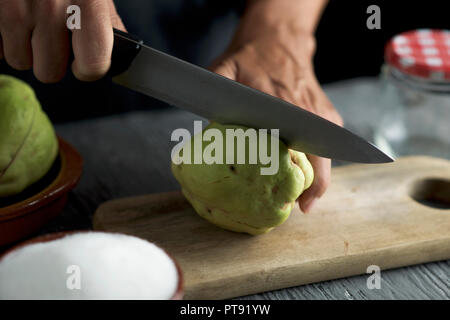 Libre d'un jeune homme de race blanche d'une coupe de fruits coing avec un couteau de cuisine, sur une planche, sur une table en bois rustique gris Banque D'Images