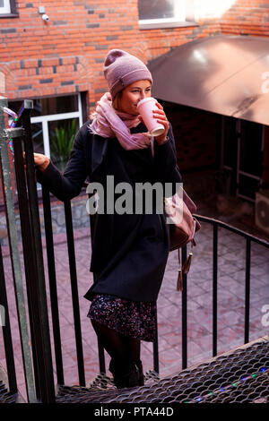 Young attractive dame en rose knitting chapeau et manteau noir pour aller boire un café . Beau sourire hipster happy woman in city street, le port c élégant Banque D'Images