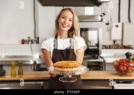 Souriante jeune femme chef cuisinier en tablier, debout à la cuisine savoureuse, montrant pie Banque D'Images