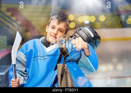 Portrait of happy Young boy, joueur de hockey sur glace, holding casque et stick, debout à côté de bardes patinoire Banque D'Images