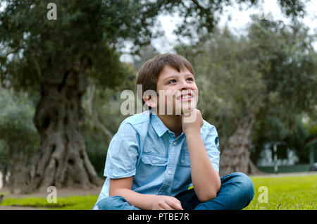 Cute little boy sitting in the park smiling pense à quelque chose Banque D'Images