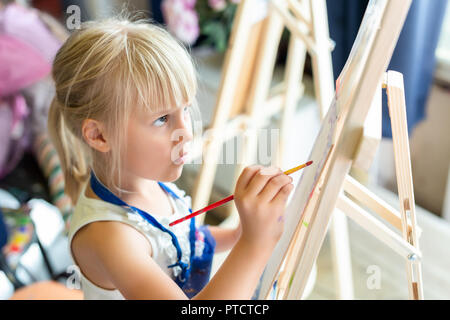 Cute blonde smiling girl painting on easel in workshop leçon à art studio. Kid holding pinceau dans la main et s'amusant à dessiner avec les peintures. Développement de l'enfant Banque D'Images