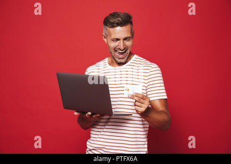 Image de l'homme barbu en t-shirt à rayures smiling while holding credit card et ordinateur portable isolé sur fond rouge Banque D'Images