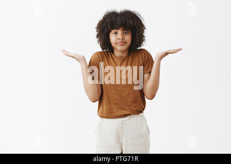 Studio shot of clueless attrayant et élégante jeune femme féminine urbaine avec afro hairstyle dans brown t-shirt faisant hausser des épaules avec les mains de côté la recherche de propagation lèvres n'ayant aucun intérêt et aucune idée Banque D'Images