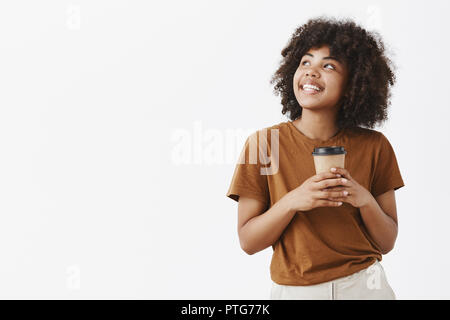 Studio shot of rêveuse romantique bonne-fille à la coiffure afro avec en brun t-shirt holding paper tasse de café et contemplant avec sourire joyeux dans le coin en haut à gauche profitant de boisson chaude et de beau temps Banque D'Images