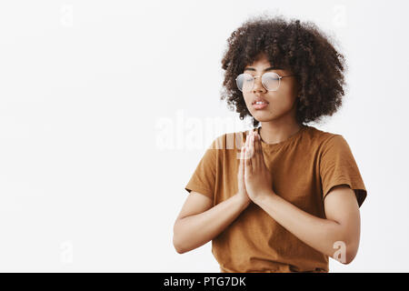 Taille plan sur l'accent calme et détendu belle jeune femme à la peau sombre dans les verres avec coiffure afro à moitié debout tourné à gauche avec les yeux fermés dans les mains ou geste namaste prier méditer Banque D'Images