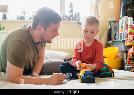 Littl garçon et de son père assis sur son lit dans la chambre à coucher et de jouer avec quelques camions de jouets. Banque D'Images