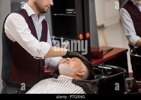 Méconnaissable coiffure tête de balayage avec la clientèle en noir serviette lavabo. Coiffure homme cheveux style de vieux client avec barbe grise. Hairstylist travaillant dans un salon de barbier. Banque D'Images