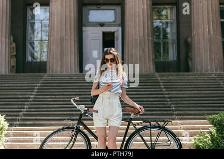 Jolie jeune fille à lunettes en utilisant smartphone pendant que debout avec vélo près d'un bel immeuble avec colonnes et escaliers Banque D'Images