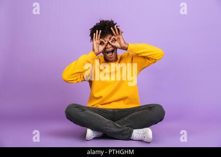 Portrait of african american man drôle assis au sol avec les jambes croisées et vous regarde dans les jumelles de décisions isolées avec les doigts au-dessus de viole Banque D'Images