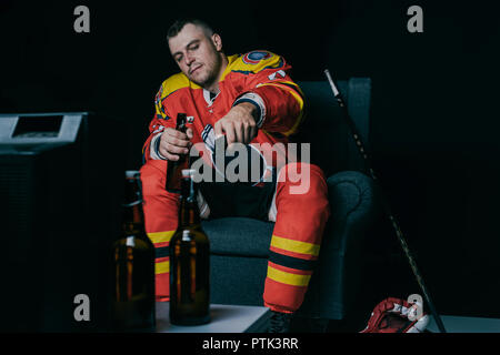 Joueur de hockey à l'aide de la télécommande et de boire de la bière tout en restant assis et regarder la télévision sur noir Banque D'Images