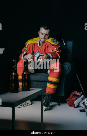 Jeune joueur de hockey à l'aide de la télécommande et de boire de la bière tout en restant assis dans un fauteuil et regarder la télé sur noir Banque D'Images