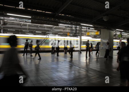 Les passagers dans une station de train : un train jaune passe rapidement dans une gare ferroviaire, en face de passagers en attente et de marcher sur la plate-forme. Banque D'Images