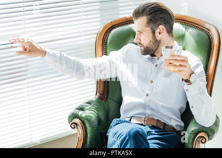 L'homme élégant barbu avec verre de whisky et assis dans un fauteuil vert et à la recherche dans la fenêtre Banque D'Images