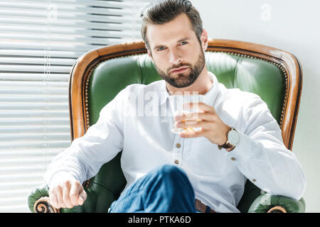 Bel homme élégant holding glass of whiskey et assis dans un fauteuil vert Banque D'Images