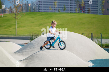 Un petit garçon jouit d'un équilibre vélo sur collines artificielles sur une aire de jeux dans un parc sur une journée ensoleillée d'été. Banque D'Images