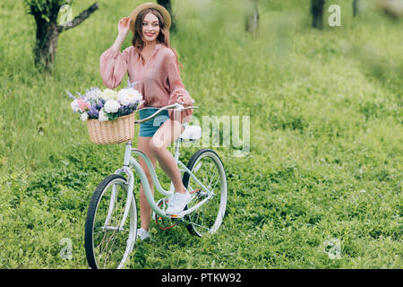 Pretty smiling woman on retro vélo avec panier en osier plein de fleurs en forêt Banque D'Images