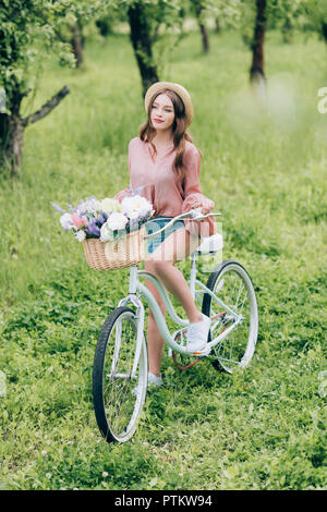 Jeune femme sur retro vélo avec panier en osier plein de fleurs en forêt Banque D'Images