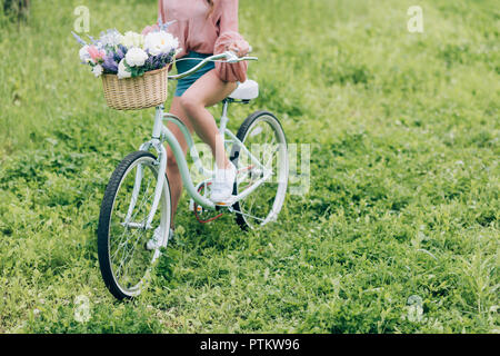 Vue partielle de la femme on retro vélo avec panier en osier plein de fleurs en forêt Banque D'Images