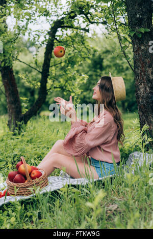 Pretty young woman in hat reposant sur couverture avec panier en osier avec des pommes dans park Banque D'Images