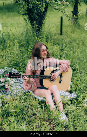 Jeune femme jouant de la guitare acoustique tout en vous reposant sur couverture avec bouquet de fleurs dans le parc Banque D'Images