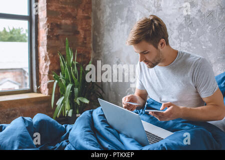 Jeune homme au lit avec carte de crédit et faire des achats en ligne sur smartphone ordinateur portable Banque D'Images