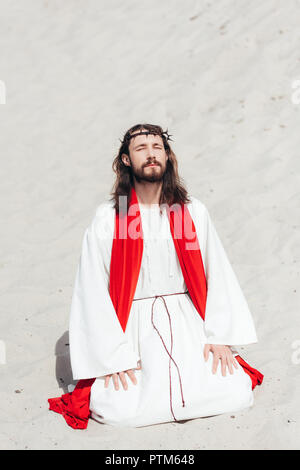 Jésus en robe, ceinture rouge et couronne d'épines debout sur les genoux avec les yeux fermés et en priant dans le désert Banque D'Images