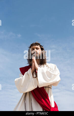 Low angle view of Jesus in robe, ceinture rouge et couronne d'épines holding rosaire et de prier avec les yeux fermés contre le ciel bleu Banque D'Images