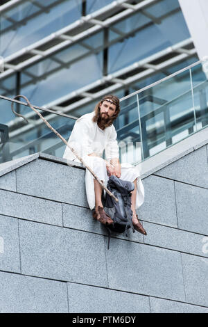 Low angle view of Jésus en robe et couronne d'épines assis avec un sac de voyage et le personnel sur le côté d'escalier Banque D'Images