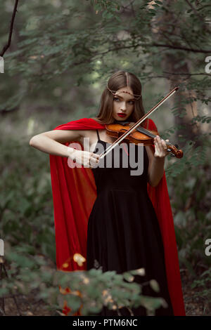 Belle fille mystique en manteau rouge jouant au violon en bois sombre Banque D'Images