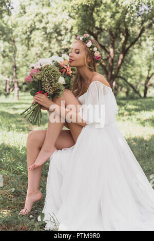 Beautiful happy young barefoot bride holding bouquet of flowers in park Banque D'Images