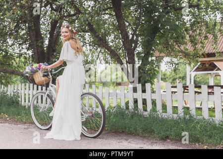 Belle jeune femme en robe de mariage riding bicycle and smiling at camera Banque D'Images