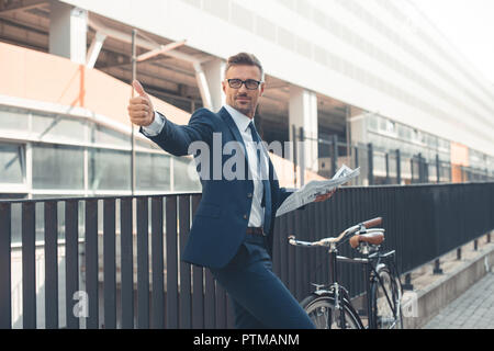 Smiling businessman holding newspaper et showing thumb up en allant à vélo sur street Banque D'Images