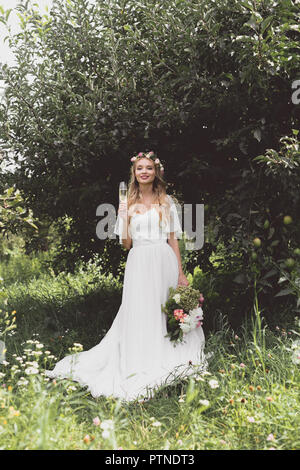 Belle jeune bride holding bouquet de mariage et le verre de champagne, smiling at camera in garden Banque D'Images