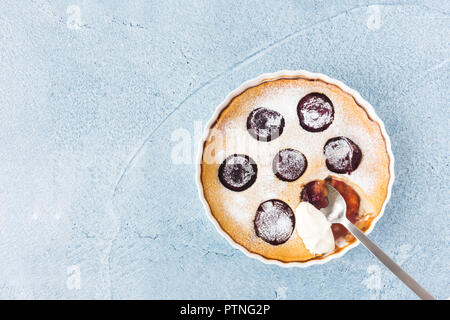 Des Clafoutis aux prunes dans une forme de céramique, avec cuillère et crème fouettée sur fond de béton bleu. Vue d'en haut. Banque D'Images