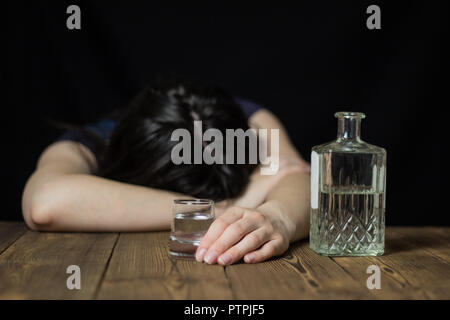 Une jeune fille ivre posé sa tête sur une table, dans sa main un verre d'alcool et une bouteille, une table en bois, un fond noir Banque D'Images