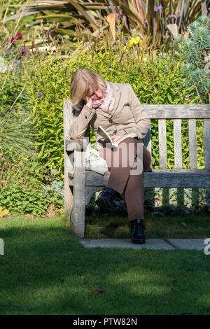 Une vieille dame d'âge moyen ou ou d'une femme assise sur un banc de parc en bois et la lecture d'un livre pendant la pause déjeuner. Banque D'Images