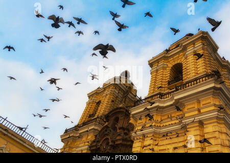 Un troupeau de pigeons volent en cercles contre un ciel bleu autour de l'église de San Francisco, à Lima, au Pérou, une cathédrale à deux tours de cloche peinte en jaune. Banque D'Images