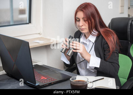 Jeune professionnel femme assise au bureau devant un ordinateur portable, en regardant à travers des jumelles, elle se concentre, à la recherche de quelque chose Banque D'Images