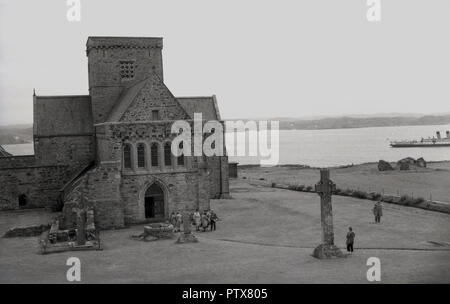 I1950s, historique, une vue de l'extérieur de la cité médiévale de l'abbaye d'Iona et environs, à l'île de Iona, Hébrides, îles de l'ouest, de l'Écosse, au Royaume-Uni. Un lieu sacré, l'un des plus anciens centres religieux chrétiens en Europe de l'Ouest et le site de pèlerinage chrétien. , 'St Martin's Cross', un historicstone cross, peut être vu dans l'avant-plan à l'extérieur de l'abbaye. Le droit à un passager steamship est mouillée. Banque D'Images