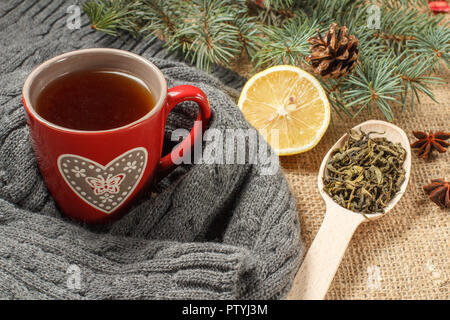 Tasse de thé rouge avec motif en forme de coeur enveloppé dans l'écharpe de laine, tranche de citron, les branches de sapins avec cône et d'anis étoilé sur le sac Banque D'Images