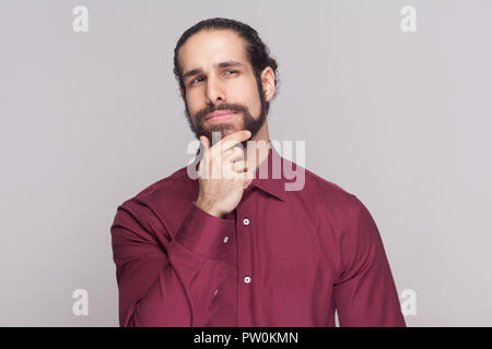 Portrait of smiling handsome man with dark recueillis des cheveux longs et la barbe en chemise rouge et permanent de toucher son menton, de la pensée et à l'hôtel. au Banque D'Images