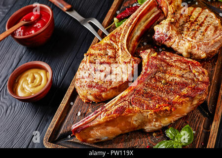 Close-up de steaks grillés ou de cow-boy tomahawk les steaks sur une vieille planche à découper en bois grossier avec fourche sur un tableau noir avec du ketchup et de la moutarde, vie Banque D'Images