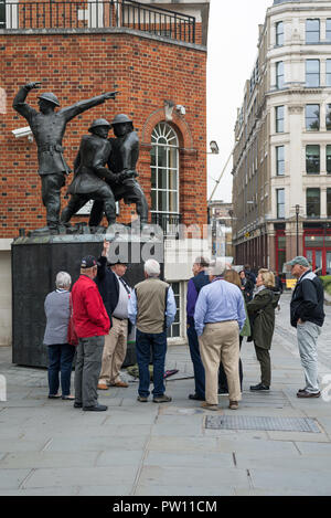 Un London tour guide avec un groupe de touristes américains à l'échelle nationale statue commémorative de pompiers dans Carter Lane Gardens, London, England, UK Banque D'Images